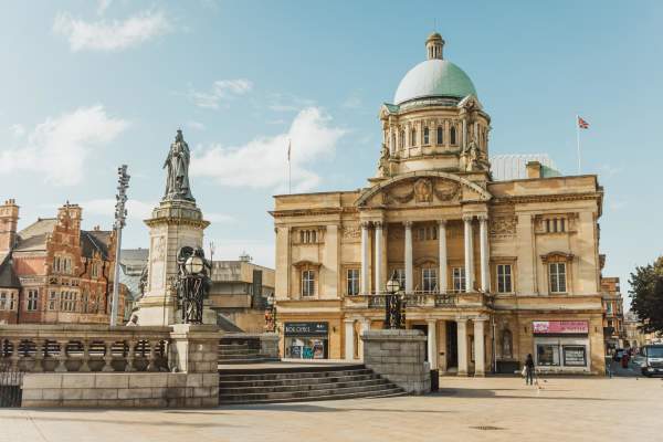 Queen Victoria Square on a sunny day. Hull City Hall is in the background, the early 20th-century Baroque Revival columns and blue copper dome look striking in the sun, The statue of Queen Victora is standing proud to the left of the image.