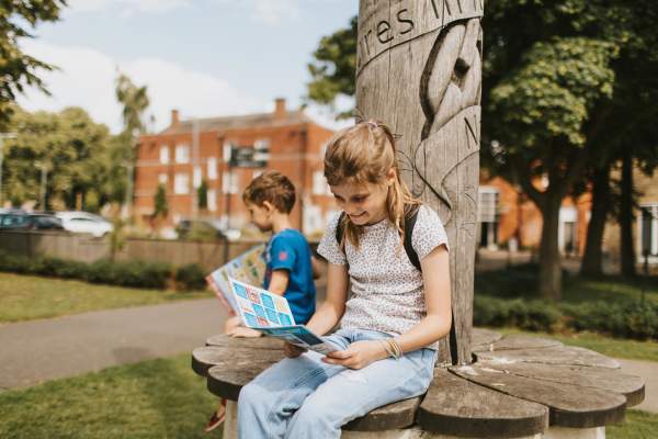 Girl looking at a treasure trail map sat on a bench with young boy in background