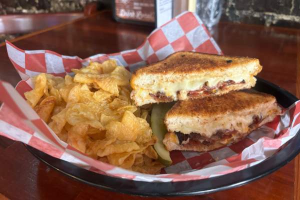 Grilled cheese with bacon, cut in half with chips and a pickle in a basket with red and white checkerboard paper