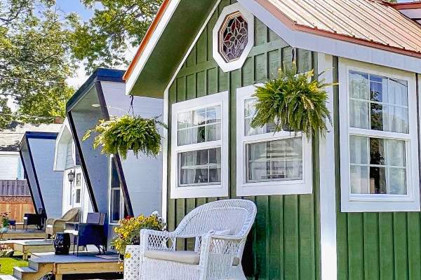 Green vertical-sided tiny house with a small front porch furnished with white wicker seating, set against a blue sky and leafy green tree.