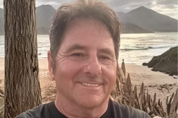 Portrait of Scott Dodson smiling on a beach with a sandcastle and hills in the background.