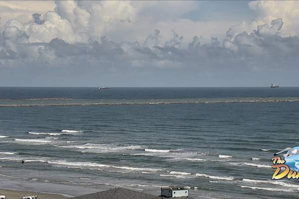 Screenshot of a live beach cam looking out on the jetties and water. In the top left hand corner is a red "Live" symbol and in the bottom right is a logo for The Dunes