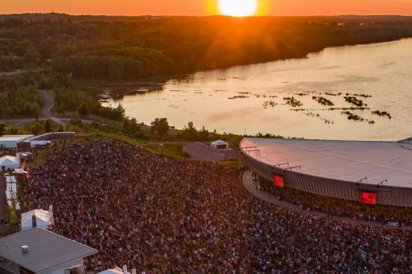 Aerial shot of Lakeview Amphitheater, large crowd in front of amphitheater set on the shores of Onondaga Lake, a fiery orange sunset can be seen in the horizon