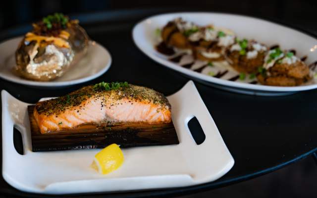 An enticing photo of a serving tray featuring salmon with a wedge of lemon, a baked potato, and fried green tomatoes drizzled with balsamic reduction plated beautifully on white plates of various sizes and shapes.