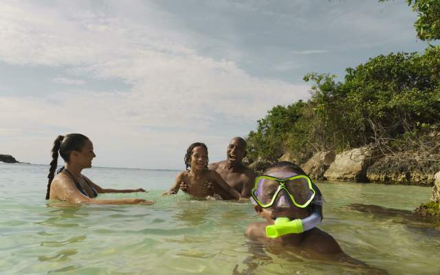 A family plays together in the water at a beach in Jamaica. In the foreground, a little boy with goggles and a snorkle looks at the camera.
