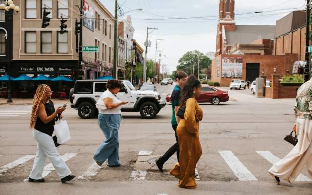 People walking across a crosswalk on a sunny day in downtown Lexington, with historic brick buildings, storefronts, and city streetlights in the background.