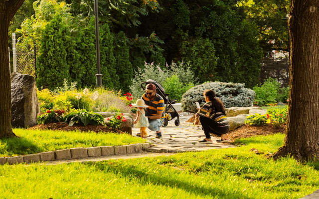 Children leaning over a stone-lined stream in a landscaped park in Providence, Rhode Island, surrounded by trees, grass, and garden plantings on a sunny day.