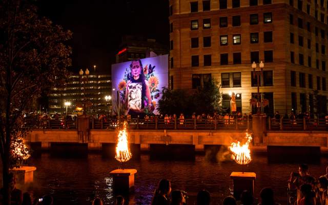 Braziers burn on the river and a mural of a young girl is illuminated in the background.