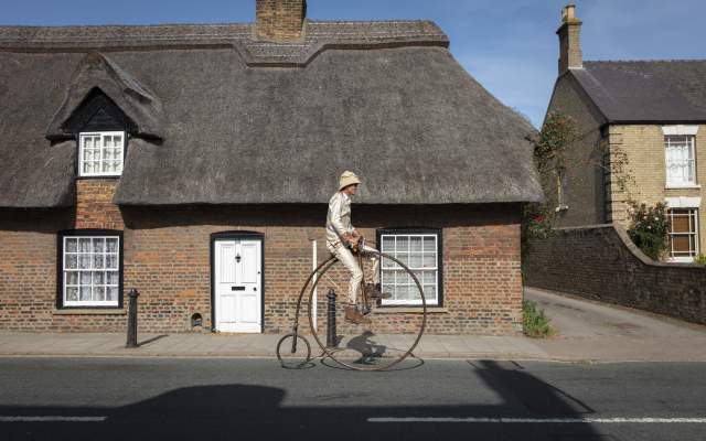 a man on a penny farthing in front of a thatched house