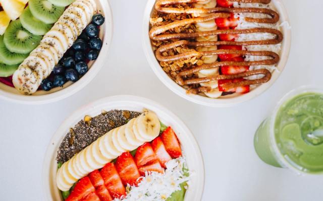 Three bowls of fruit and one green smoothie on a white table.