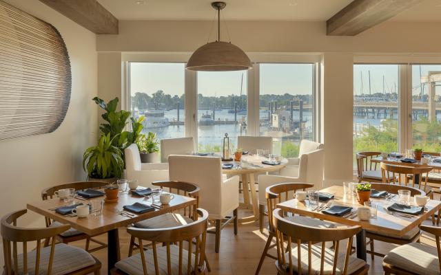 A dining area with multiple tables set for meals, large windows, and a view of the water.