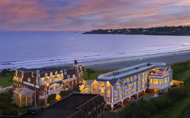A large hotel building with lit windows sits on a beachfront property overlooking the ocean at sunset.
