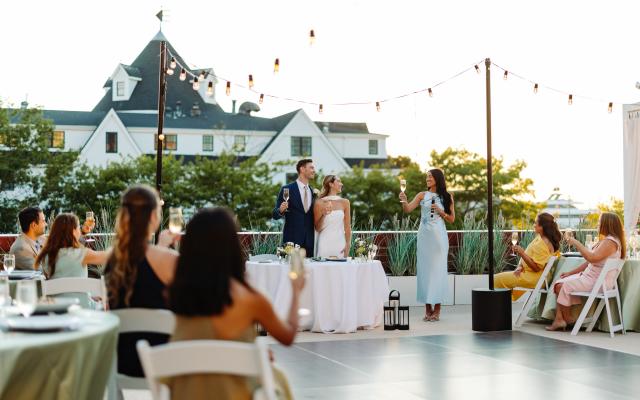 A wedding reception with guests seated at tables and a bride and groom standing at a podium in an outdoor event space with string lights and a large white building in the background.