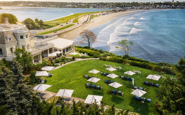 A large white building with a grassy area and umbrellas in front of a beach and ocean.