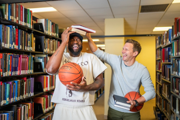 Wade & Diener in a library laughing, holding basketballs and books. Deiener holding books on Wade's head.