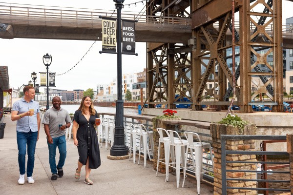 Three people walk along the Milwaukee Riverwalk outside Lakefront Brewery, holding beers and smiling. They pass under an industrial bridge structure with banners reading “Cold Beer in Here” and “Beer. Food. Beer.” while white bar stools and flower planters line the railing overlooking the river.