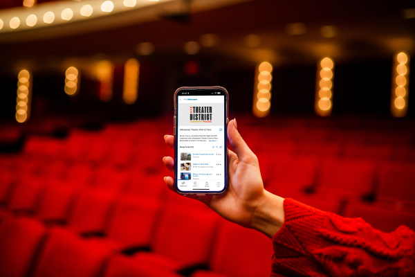 Hand holding up cellphone displaying homescreen of Milwaukee Theater District Pass in front of blurry theater background