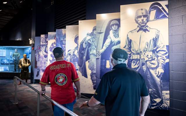 Two visitors walk through a military history exhibit lined with large blue-toned panels of paratroopers and nearby display cases at Fayetteville's ASOM.