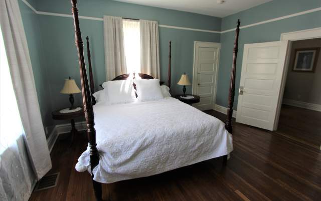 Guest bedroom at MacPherson House Bed & Breakfast featuring a four-poster bed with white linens, hardwood floors, soft blue walls, and natural light from tall windows.