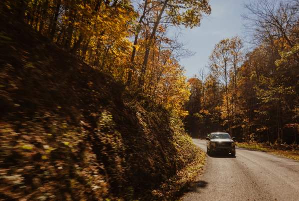 A truck drives along a paved road during a sunny day with fall foliage along the roadside.