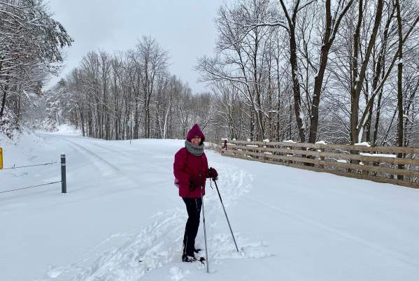 A woman is bundled up in winter gear and wearing corss country skis with poles in hand at a snow-covered trail head.
