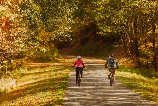 A couple ride bike on a gravel path surrounded trees with fall colored leaves of yellows, oranges, and reds.