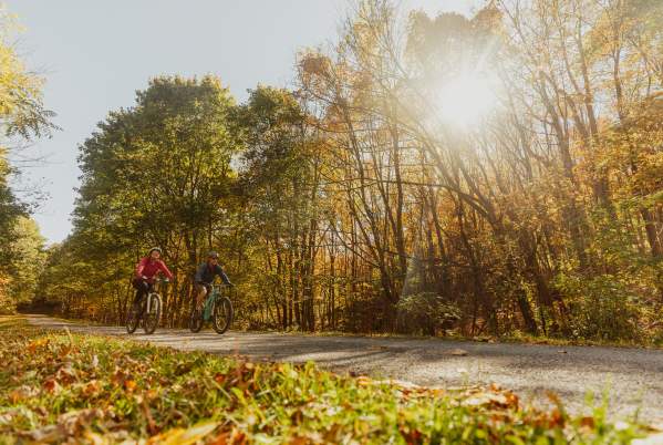 A man and a woman are each on their own bike. peddling along the Great Allegheny Passage in the fall.