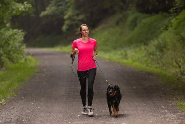 A woman in a bright pink shirt and black pants walks a black and brown, medium-sized dog on a gravel through a wooded area.