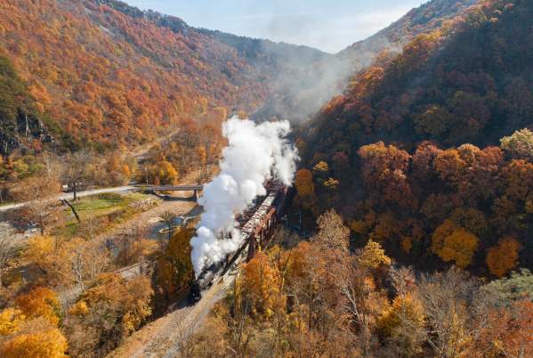 A steam train travels through a mountain valley surrounded by vibrant autumn foliage.