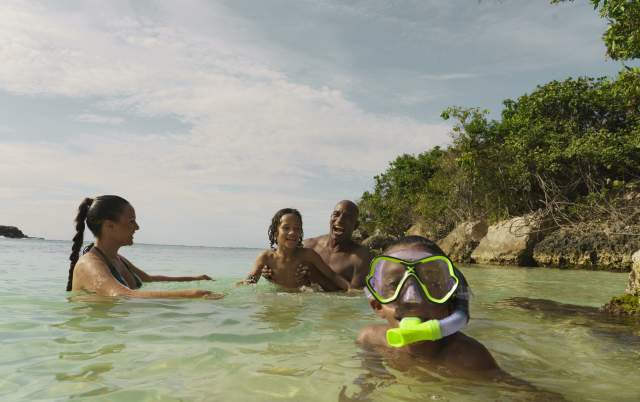 A family plays together in the water at a beach in Jamaica. In the foreground, a little boy with goggles and a snorkle looks at the camera.