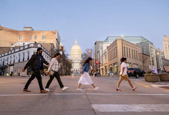 A family of four visiting Madison, WI walks across the street with the Wisconsin State Capitol and art museum in the background
