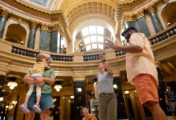 A family explores a grand, historic building with ornate architecture. They engage in conversation, surrounded by marble columns and large windows.