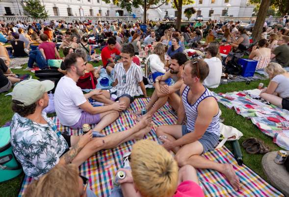 A lively outdoor gathering on a grassy area, with groups of people sitting on blankets, enjoying drinks and conversation under trees.