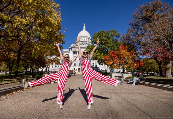 Two women wearing red and white striped overalls posing in front of the Wisconsin State Capitol building.