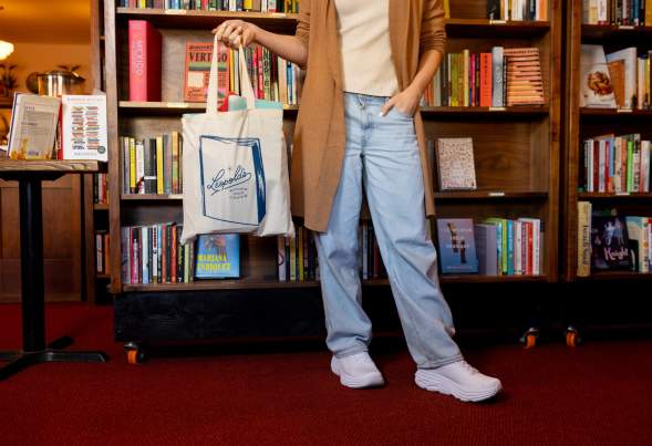 A photo from the chest down of a person holding a Leopolds Bookstore tote bag while standing in front of a bookshelf.