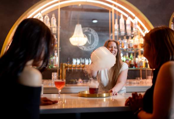 A bartender serves a cocktail with smoke effects at a stylish bar, while two patrons watch, enjoying the vibrant atmosphere.