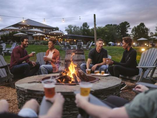 Group of friends enjoying craft beer at Lost Barrel Brewery around a firepit with scenic outdoor background and hanging lights at dusk