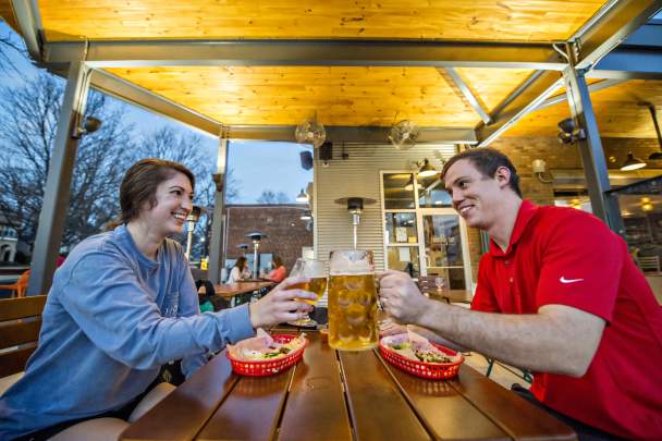 Woman and man sitting at an outdoor wooden table toasting glasses of beer