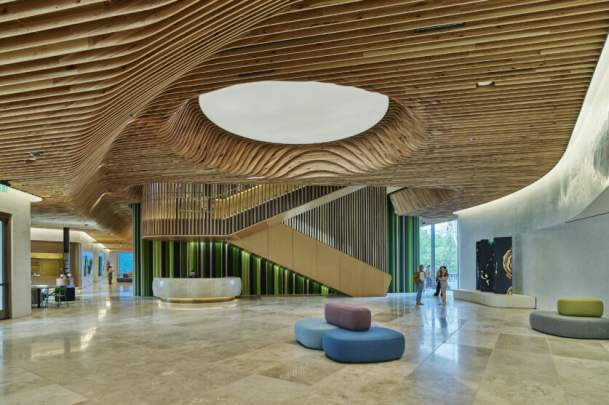 A grand, modern lobby featuring a wavy slatted wood ceiling with a circular cutout, a large wooden staircase, and a curved reception desk with a green textured front. Light-colored stone floors, colorful ottomans, and large windows with people and trees complete the scene.