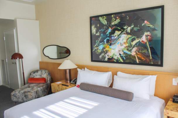 A cozy hotel room featuring a queen bed with white linens, a colorful abstract painting, a lamp, and a patterned armchair.