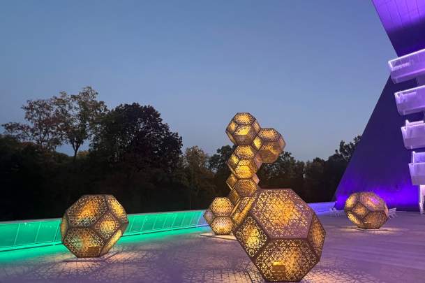 Illuminated geometric sculptures in a modern outdoor space at dusk, surrounded by trees and vibrant lighting, creating a captivating atmosphere.