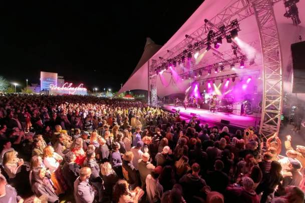 The Roots perform on a brightly lit stage under a white canopy at Momentary Green in Bentonville, Arkansas, before a large, dark crowd at night. Pink and white spotlights illuminate the band.
