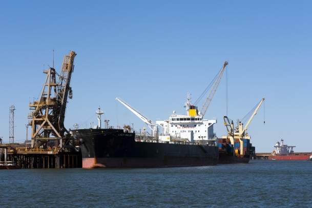 An Iron ore carrier in the Port of Port Hedland, seen from the water with a tug boat along side and a crane on land.