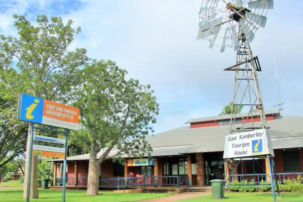 Image depicts the Kununurra Visitor Centre from a front, slightly off centre angle. In the foreground stands an old windmill with the sign on and some boab trees and a standing sign on the opposite side of the footpath. The building is brick with a tin roof.