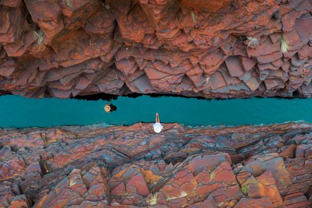 Overhead shot of two people in a narrow section of Weano Gorge, Karijini National Park. One is walking along the gorge floor, through water, and one is relaxing on the rocks.