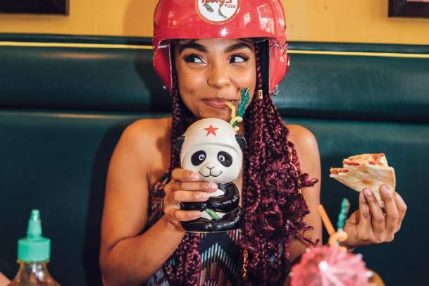 Woman dining at Fong's Pizza wearing a Fong's helmet, drinking out of a panda cup, and eating pizza