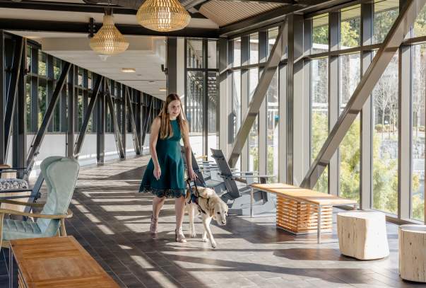 A woman in a green dress walks with a service dog. She is walking along a bridge inside a building with sunlight streaming in from the many windows.