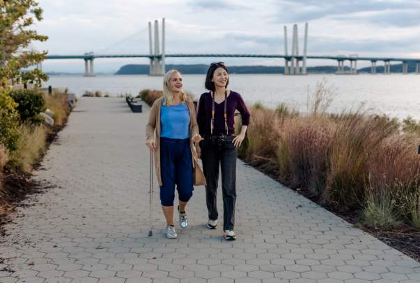 Two people are walking along the Riverwalk in Tarrytown. The Gov. Mario Cuomo Bridge is in the background.