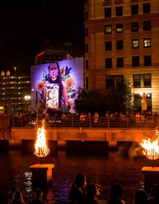 Braziers burn on the river and a mural of a young girl is illuminated in the background.