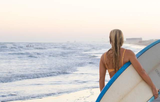 A girl carrying a surfboard on her side walks down the beach toward the pier.